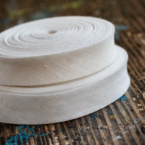 May include: Two rolls of white cotton twill tape, a type of woven fabric ribbon, are stacked on top of each other on a wooden surface.
