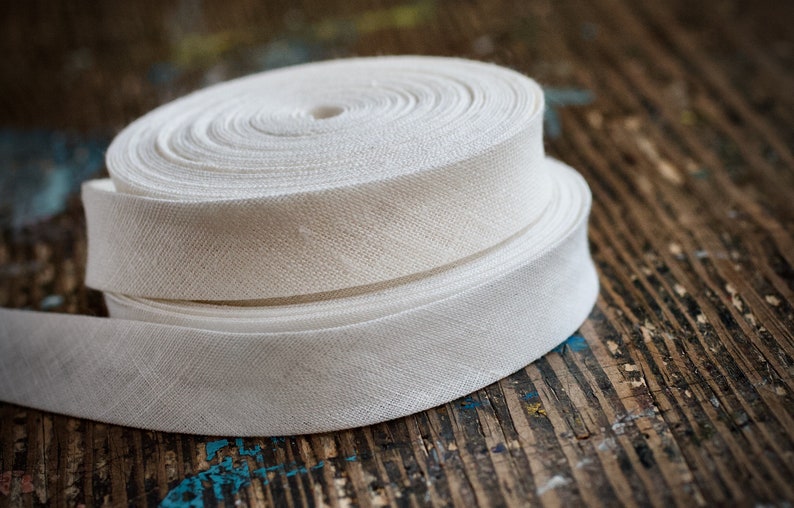 May include: Two rolls of white cotton twill tape, a type of woven fabric ribbon, are stacked on top of each other on a wooden surface.
