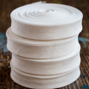 May include: Four spools of white cotton twill tape stacked on top of each other. The tape is rolled up and has a subtle herringbone pattern.