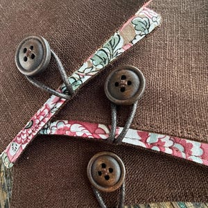 May include: Close-up of a brown fabric with three wooden buttons. The buttons are attached with dark thread loops. Floral patterned fabric trim in pink, green, and white accents the brown fabric.