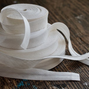 May include: A stack of white cotton bias tape rolls on a wooden surface. The tape is folded and ready for sewing projects.