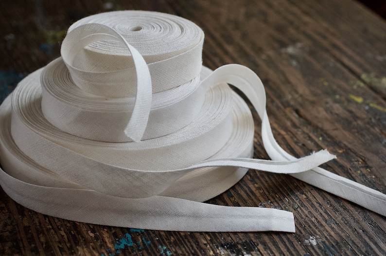 May include: A stack of white cotton bias tape rolls on a wooden surface. The tape is folded and ready for sewing projects.