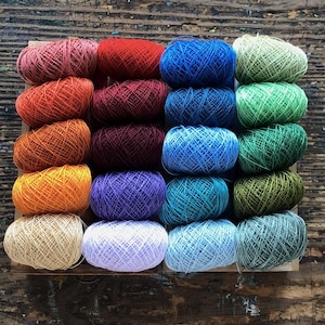 May include: A collection of 20 colorful yarn spools in a wooden box. The spools are arranged in rows of four, with each row featuring a different color. The colors include red, orange, yellow, green, blue, purple, and white.