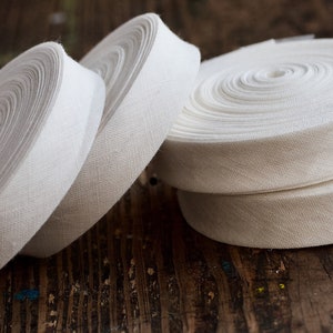 May include: Four spools of white cotton twill tape, rolled and stacked on a wooden surface.