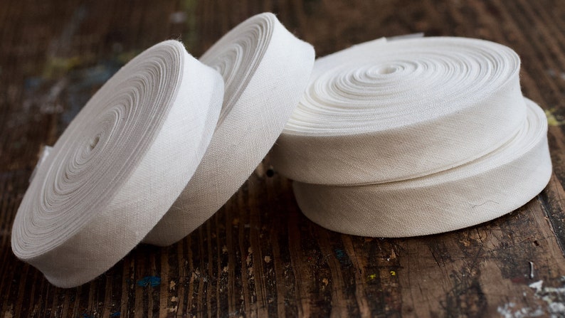 May include: Four spools of white cotton twill tape, rolled and stacked on a wooden surface.
