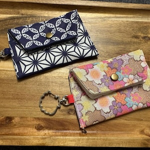 May include: Two small fabric wallets with keychains on a wooden tray. One is navy blue with white geometric and floral patterns. The other is floral with pink, orange, and yellow flowers.