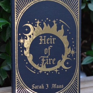 May include: A black hardcover book with gold foil lettering and a decorative design. The title is "Heir of Fire" by Sarah J. Maas.