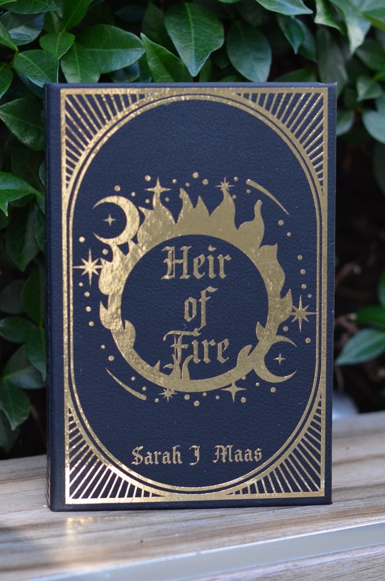 May include: A black hardcover book with gold foil lettering and a decorative design. The title is "Heir of Fire" by Sarah J. Maas.