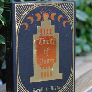May include: A black hardcover book with gold accents and a gold and orange illustration of a tower. The title of the book is "Tower of Dawn" by Sarah J. Maas.