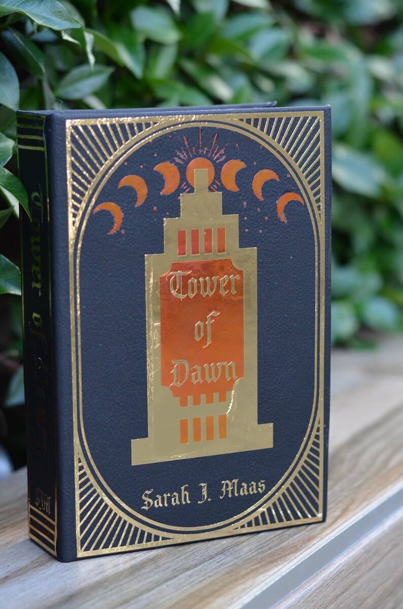 May include: A black hardcover book with gold accents and a gold and orange illustration of a tower. The title of the book is "Tower of Dawn" by Sarah J. Maas.