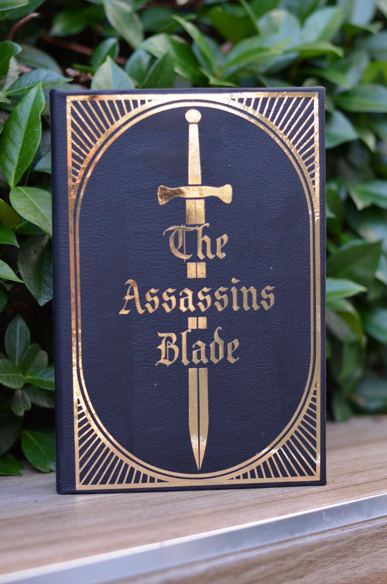 May include: A black book with a gold-embossed design. The cover features a sword with a gold blade and the title "The Assassins Blade".