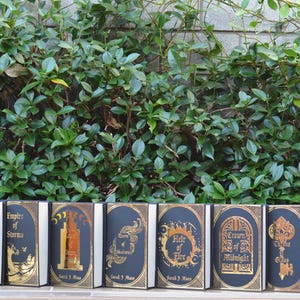 May include: A set of eight black hardcover books with gold foil lettering and illustrations. The titles are "Kingdom of Ash", "Empire of Storms", "Tower of Dawn", "A Court of Thorns and Roses", "Heir of Fire", "Crown of Midnight", "A Court of Mist and Fury", and "The Assassin's Blade".