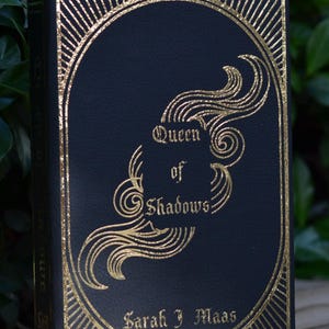 May include: A black hardcover book with gold foil lettering and decorative designs. The title is "Queen of Shadows" by Sarah J. Maas.