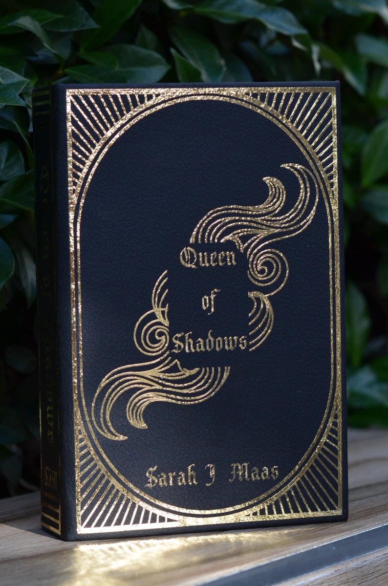 May include: A black hardcover book with gold foil lettering and decorative designs. The title is "Queen of Shadows" by Sarah J. Maas.