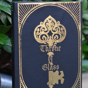 May include: A black book with a gold embossed design of a key with the words "Throne of Glass" on the cover.