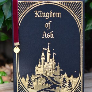 May include: A black leather-bound book with gold accents and a red ribbon bookmark. The title "Kingdom of Ash" is written in gold lettering on the front cover. A gold illustration of a castle on a hill with trees is featured on the cover.