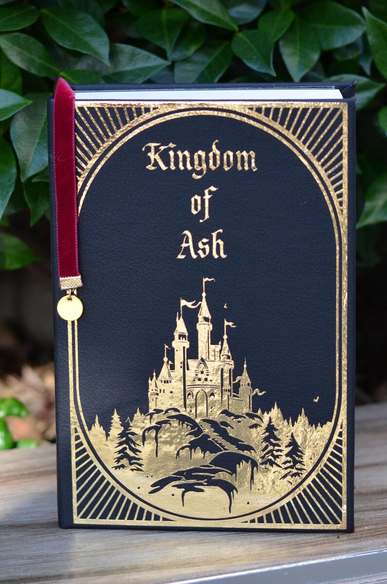 May include: A black leather-bound book with gold accents and a red ribbon bookmark. The title "Kingdom of Ash" is written in gold lettering on the front cover. A gold illustration of a castle on a hill with trees is featured on the cover.