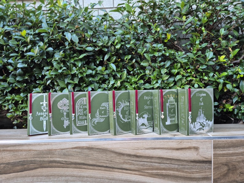 May include: A set of eight green hardcover books with silver and white illustrations and red ribbon bookmarks. The titles of the books are "The Assassin's Blade", "The Thief", "Crown of Midnight", "Queen of Shadows", "Heir of Fire", "Empire of Storms", "Tower of Dawn", and "Kingdom of Ash".