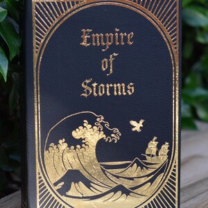 May include: A black book with gold lettering and a gold illustration of a wave and a ship. The title of the book is "Empire of Storms".