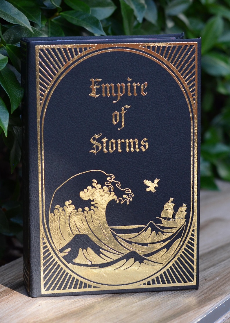 May include: A black book with gold lettering and a gold illustration of a wave and a ship. The title of the book is "Empire of Storms".