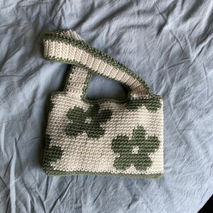 Puede incluir: Un bolso de crochet con un patr&oacute;n floral verde y blanco. El bolso tiene una sola correa y est&aacute; hecho con un hilo grueso.