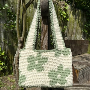 Puede incluir: Un bolso de crochet con un patr&oacute;n floral verde y blanco. El bolso tiene una correa larga y est&aacute; colgado de un poste de madera.