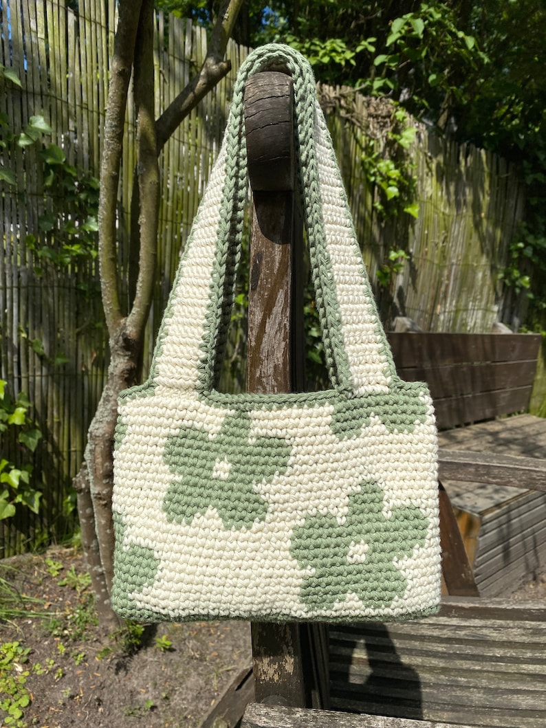 Puede incluir: Un bolso de crochet con un patr&oacute;n floral verde y blanco. El bolso tiene una correa larga y est&aacute; colgado de un poste de madera.