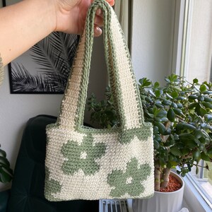 Puede incluir: Un peque&ntilde;o bolso de crochet con un patr&oacute;n floral verde y blanco. El bolso tiene dos asas y est&aacute; hecho con un hilo grueso.