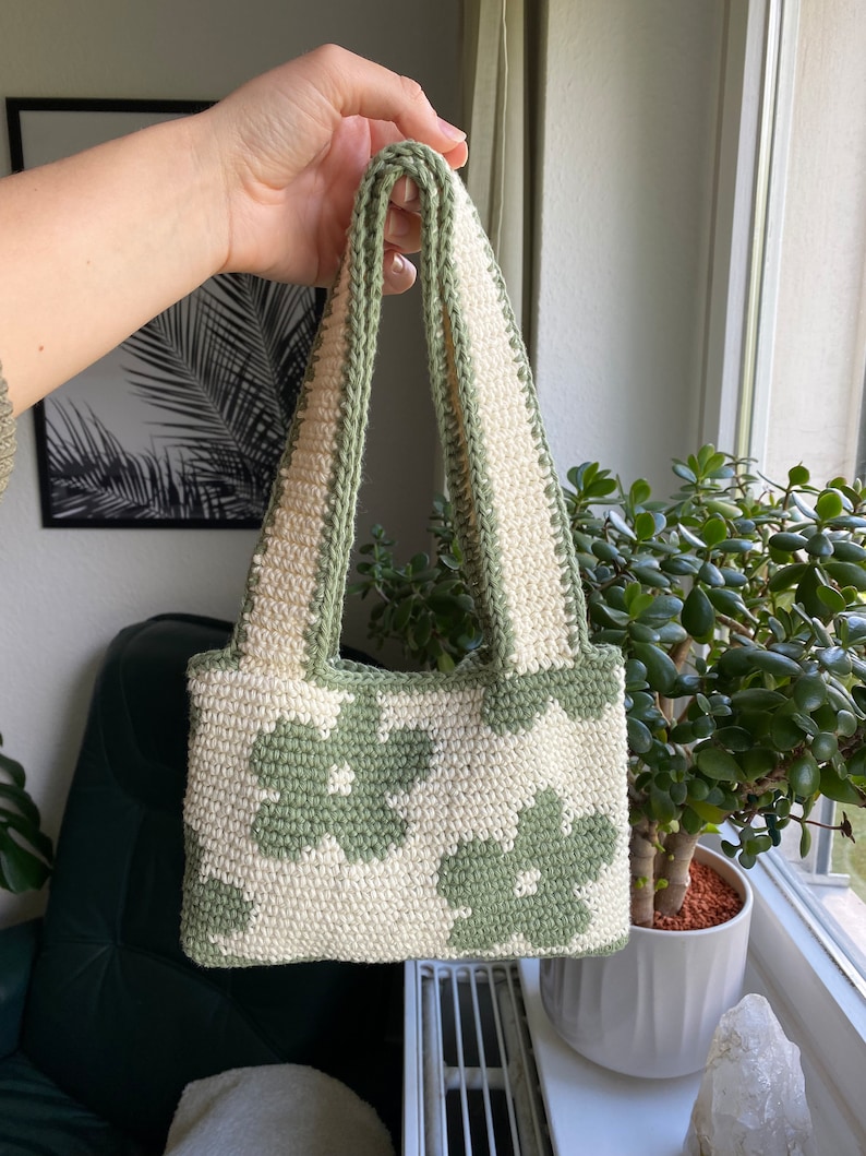 Puede incluir: Un peque&ntilde;o bolso de crochet con un patr&oacute;n floral verde y blanco. El bolso tiene dos asas y est&aacute; hecho con un hilo grueso.