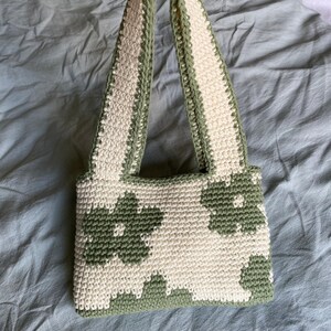 Puede incluir: Un bolso de crochet con un patr&oacute;n floral verde y blanco. El bolso tiene dos asas y est&aacute; hecho con un hilo texturizado.