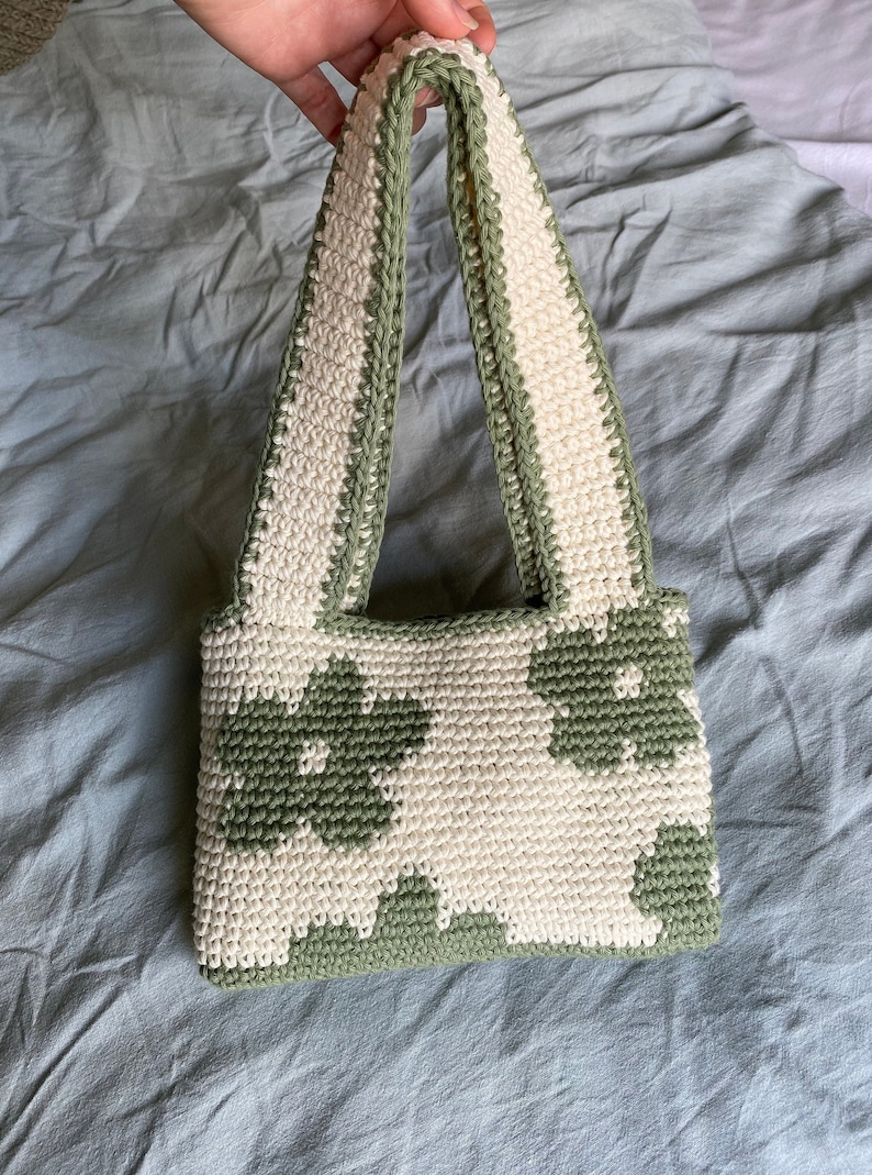 Puede incluir: Un bolso de crochet con un patr&oacute;n floral verde y blanco. El bolso tiene dos asas y est&aacute; hecho con un hilo texturizado.