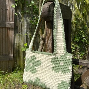 Puede incluir: Un bolso de crochet con un patr&oacute;n geom&eacute;trico verde y blanco. El bolso tiene una correa larga y est&aacute; colgado de una silla de madera.