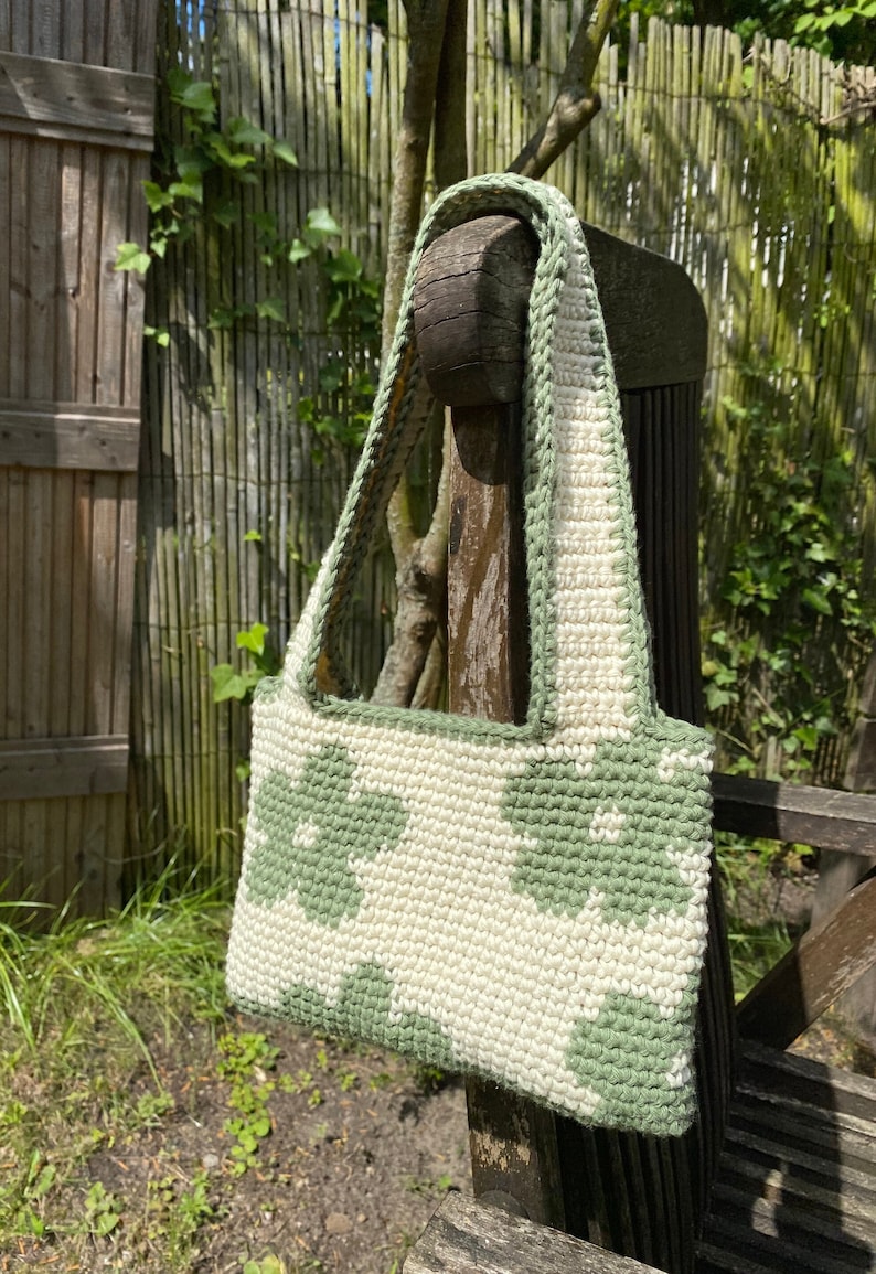 Puede incluir: Un bolso de crochet con un patr&oacute;n geom&eacute;trico verde y blanco. El bolso tiene una correa larga y est&aacute; colgado de una silla de madera.