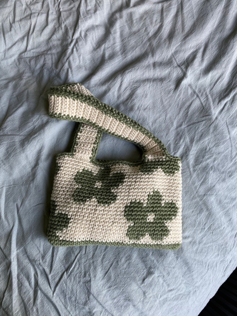 Puede incluir: Un bolso de crochet con un patr&oacute;n floral verde y blanco. El bolso tiene una sola correa y est&aacute; hecho con un hilo grueso.