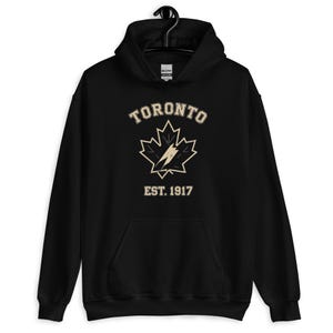 May include: Black hoodie featuring the word "TORONTO" in a vintage font above a maple leaf design with a lightning bolt. The text "EST. 1917" is below the maple leaf. The sweatshirt has a front pocket and a drawstring hood.