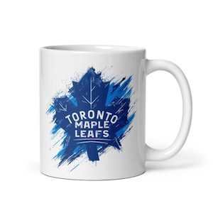 May include: White ceramic mug with the Toronto Maple Leafs logo. The logo is a blue maple leaf with the team name in white text. The design has a distressed, brushstroke effect.
