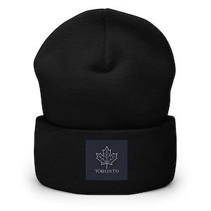 May include: Black beanie hat with a turned-up brim. A rectangular patch on the front features a white maple leaf design and the word "TORONTO". The hat is made of a soft, knitted fabric.