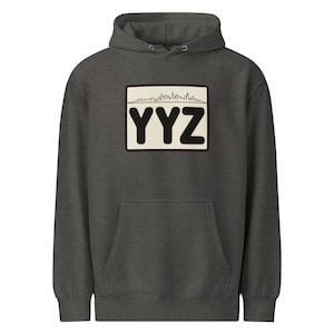 Può includere: Felpa con cappuccio grigio scuro con tasca frontale e cappuccio con coulisse. La felpa presenta una toppa rettangolare con le lettere "YYZ" in nero, uno sfondo color crema e una grafica skyline.