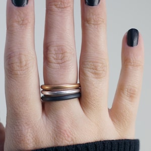 May include: A hand wearing three minimalist rings: gold, silver, and black. The rings are stacked on one finger. The nails are painted black. The person is wearing a black sweater.