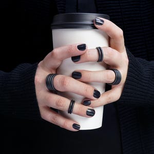 May include: A person holding a white coffee cup with a black lid, wearing multiple black rings. The person is wearing a black sweater and has black nail polish.