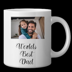 Pode incluir: Caneca de cerâmica branca com uma fotografia de um homem e uma criança, e as palavras "Worlds Best Dad" em escrita preta. A caneca tem uma alça curva e está sobre um fundo preto.