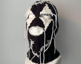 Puede incluir: Una balaclava de crochet negra y blanca con cuerdas blancas que cuelgan. La balaclava tiene un diseño en forma de estrella en la parte delantera.