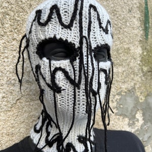 May include: A white knitted balaclava with black abstract design elements. The black design appears to drip down the face and neck. The eye and mouth openings are black.