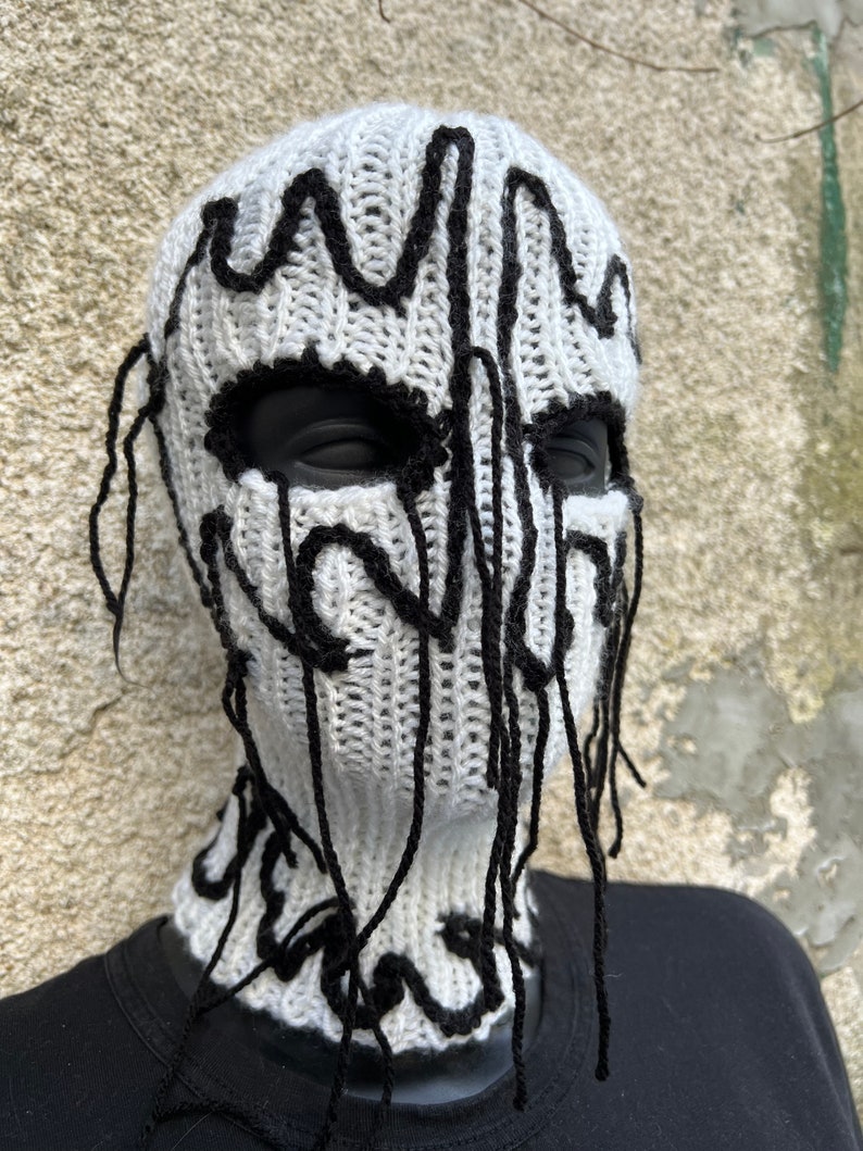May include: A white knitted balaclava with black abstract design elements. The black design appears to drip down the face and neck. The eye and mouth openings are black.