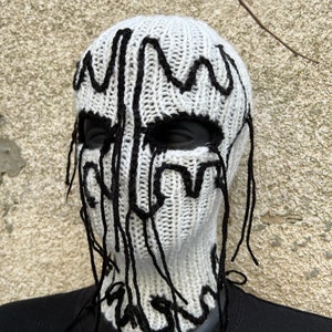 May include: White knitted balaclava with black decorative accents. The balaclava features black, dripped-style designs around the eyes and down the front. The neck is covered, and the balaclava is displayed on a black mannequin.
