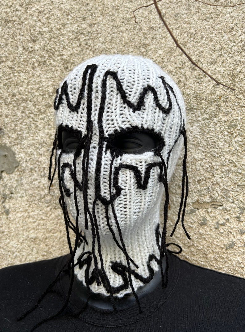May include: White knitted balaclava with black decorative accents. The balaclava features black, dripped-style designs around the eyes and down the front. The neck is covered, and the balaclava is displayed on a black mannequin.