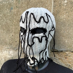 May include: A white knitted balaclava with black, dripped-style accents around the eyes and top. The balaclava is displayed on a black mannequin head. The design gives the appearance of a melting face.