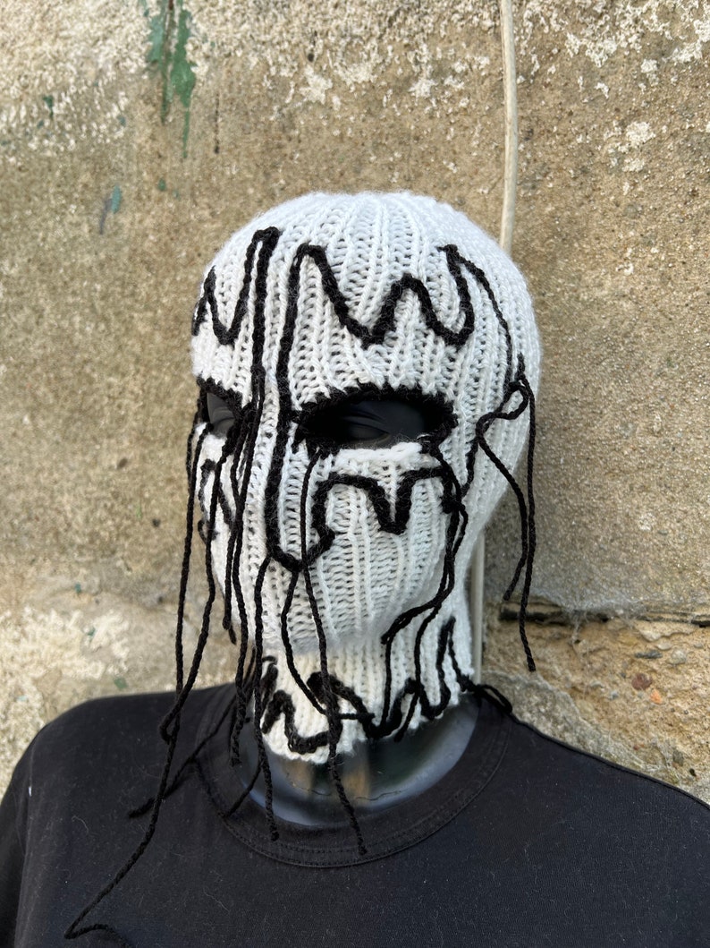 May include: A white knitted balaclava with black, dripped-style accents around the eyes and top. The balaclava is displayed on a black mannequin head. The design gives the appearance of a melting face.