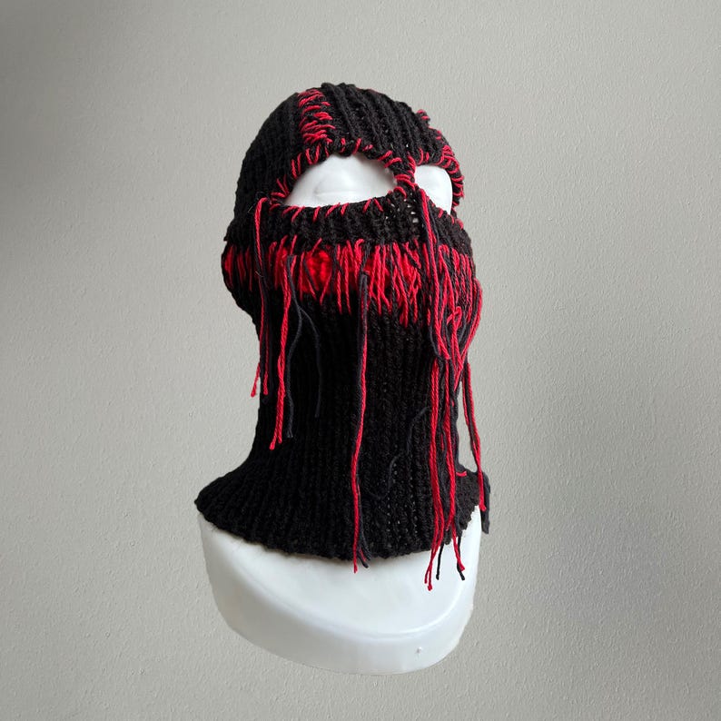 May include: Black knitted balaclava with red fringe detail. The balaclava has a cut-out for the eyes.