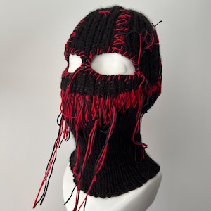 May include: Black knitted balaclava with red stitching and frayed edges. The balaclava has eye holes and covers the head and neck.